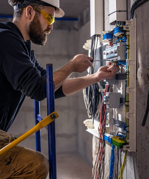 A male electrician works in a switchboard with an electrical connecting cable, connects the equipment with tools.